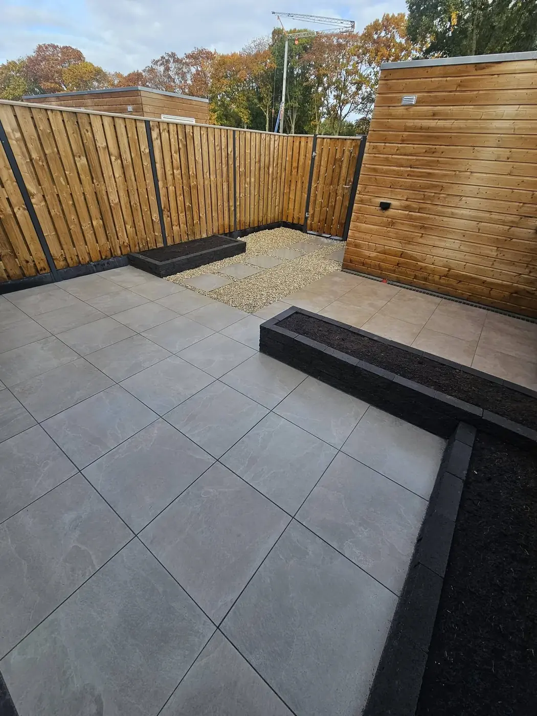 Achtertuin met keramisch terras, borders met stapelblokken, grindpad met stapstenen en een houten schutting
