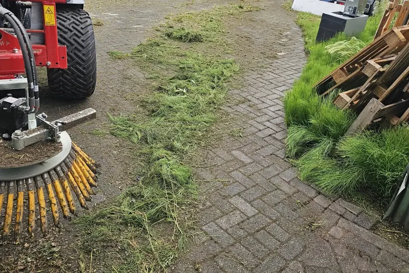 Machinaal onkruid borstelen van een klinkerpad, met het losgeborstelde gras ernaast